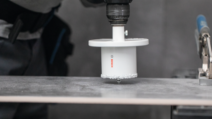 Прикључак за тестеру за рупе буши кроз керамичке плочице на радном столу.