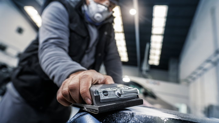 شخص يرتدي معدات السلامة يقوم بصنفرة هيكل السيارة في ورشة عمل.