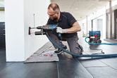 A person wearing safety equipment drills into a wall with a dust extraction attachment.