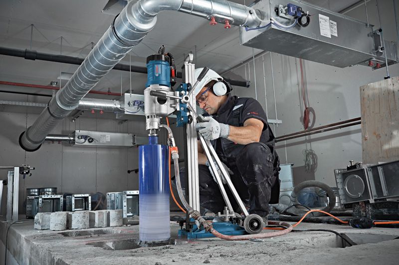 A person wearing safety equipment operates a core drill on a concrete floor.