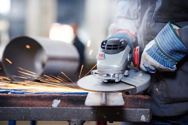 Worker wearing safety equipment grinds metal, producing sparks on a workbench.