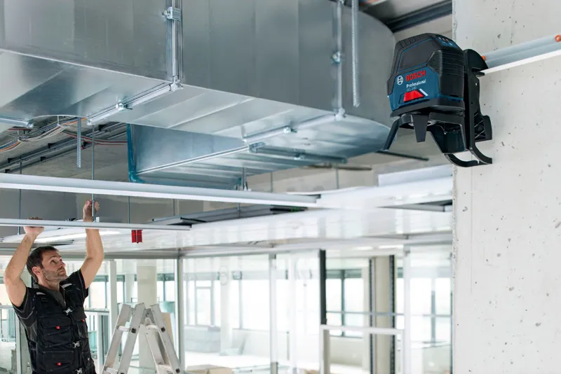 A person adjusts ceiling fixtures near a mounted laser leveling tool in an industrial setting.