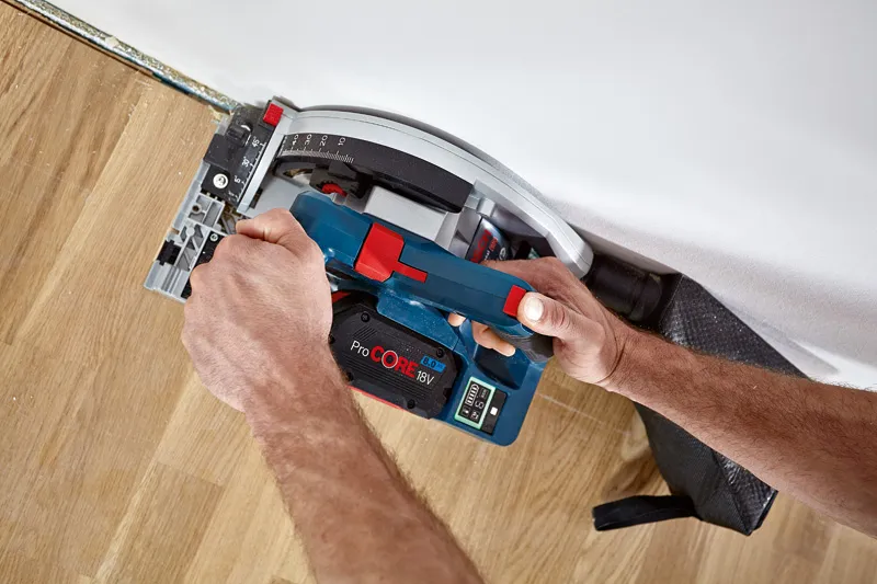 A person operates a cordless circular saw to cut wood flooring near a wall.