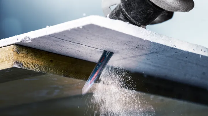 A reciprocating saw cuts through a thick concrete board, creating dust.