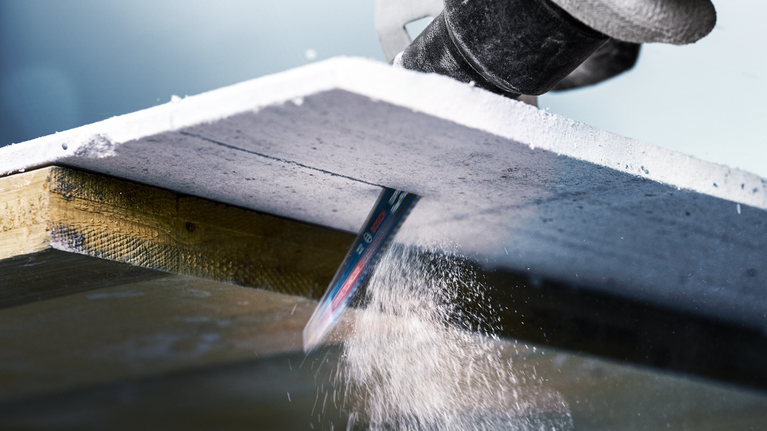 A reciprocating saw cuts through a thick concrete board, creating dust.