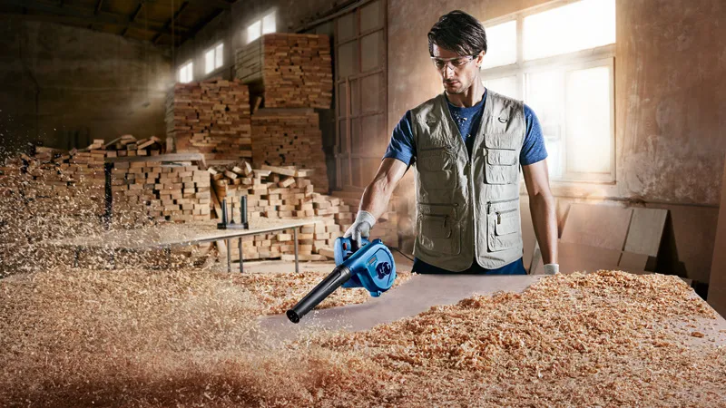A person wearing safety equipment uses a blower to clear wood shavings from a workbench.