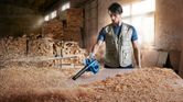 A person wearing safety equipment uses a blower to clear wood shavings from a workbench.