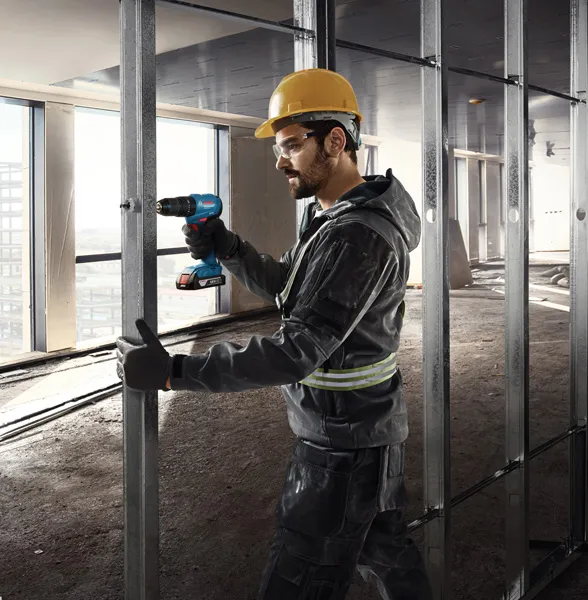 A person wearing safety equipment drills into a metal stud in a building under construction.
