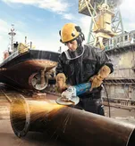 A person wearing safety equipment grinds a large metal pipe at a shipyard.