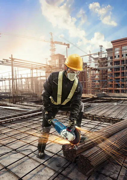 A person wearing safety equipment uses an angle grinder to cut steel rods at a construction site.