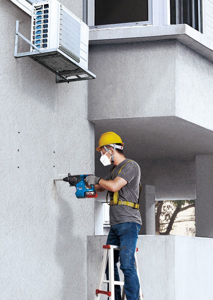 A person wearing safety equipment drills into a wall beneath an air conditioning unit.