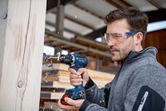 Person wearing safety equipment drills into a wooden beam in a workshop.