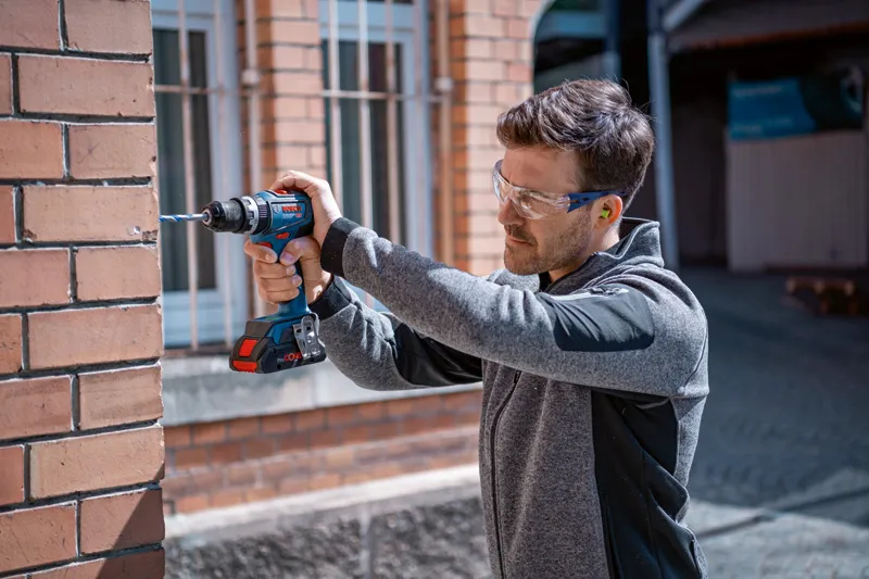 A person wearing safety equipment drills into a brick wall with a cordless power drill.