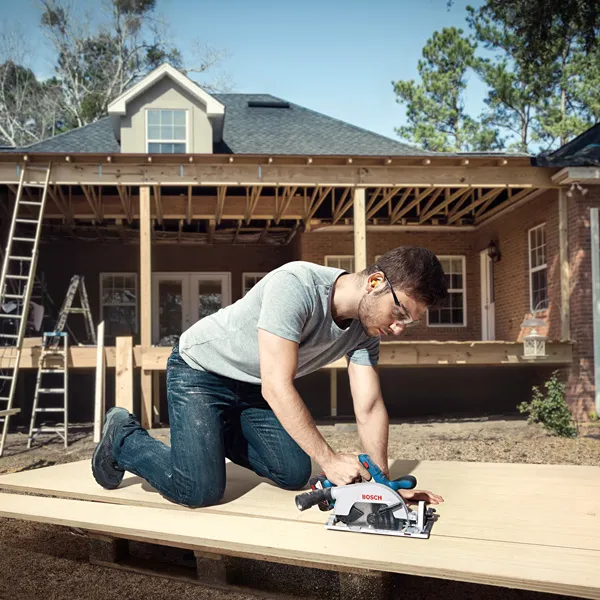 A person wearing safety equipment cuts wood with a cordless circular saw at a house construction site.