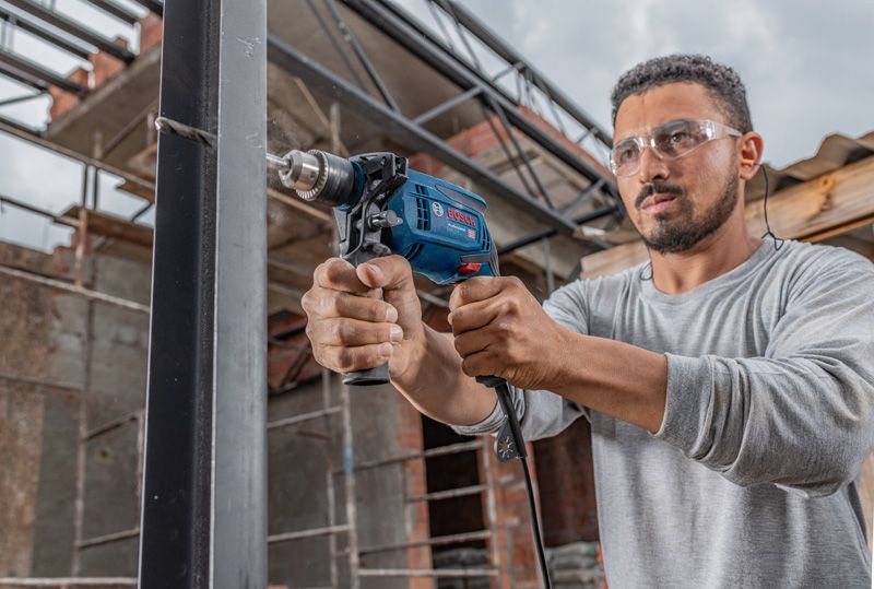 Person wearing safety equipment drills into a metal beam at a construction site.