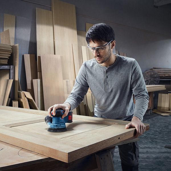 Person wearing safety equipment sands a wooden door with a power sander.