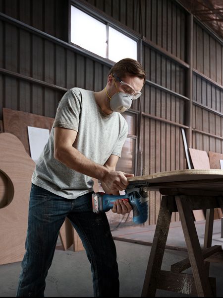 A person wearing safety equipment uses a power saw to cut wood in a workshop.