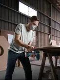 A person wearing safety equipment uses a power saw to cut wood in a workshop.
