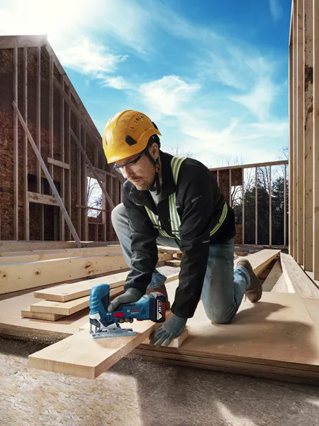 A person wearing safety equipment uses a cordless jigsaw to cut wooden planks at a construction site.