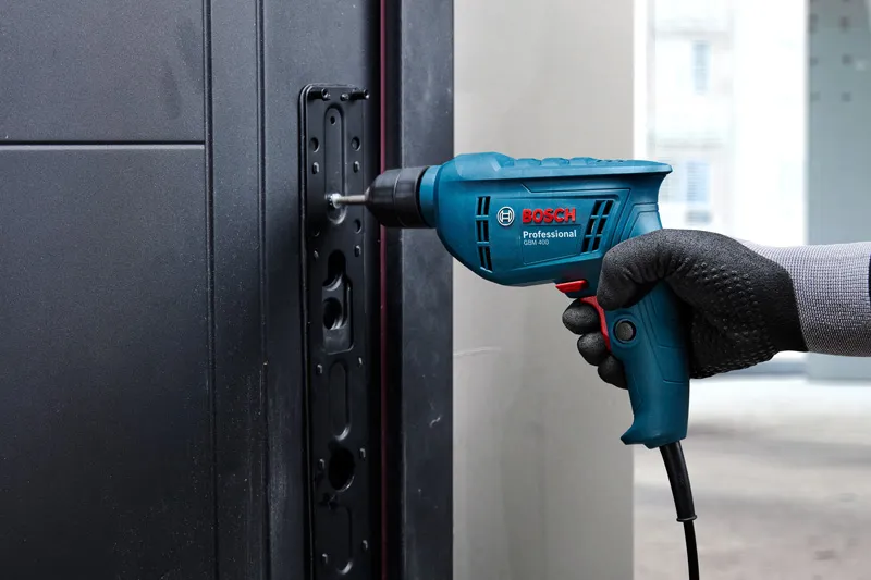 Person wearing safety equipment uses a power drill to fasten screws into a metal doorframe.