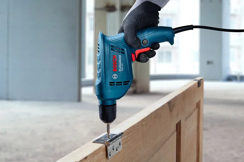 A person wearing safety equipment uses a power drill to fasten a hinge to a wooden door.
