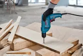 A person wearing safety equipment drills a hole in plywood on a workbench.