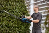 Person wearing safety equipment trims a bush outdoors using a cordless hedge trimmer.