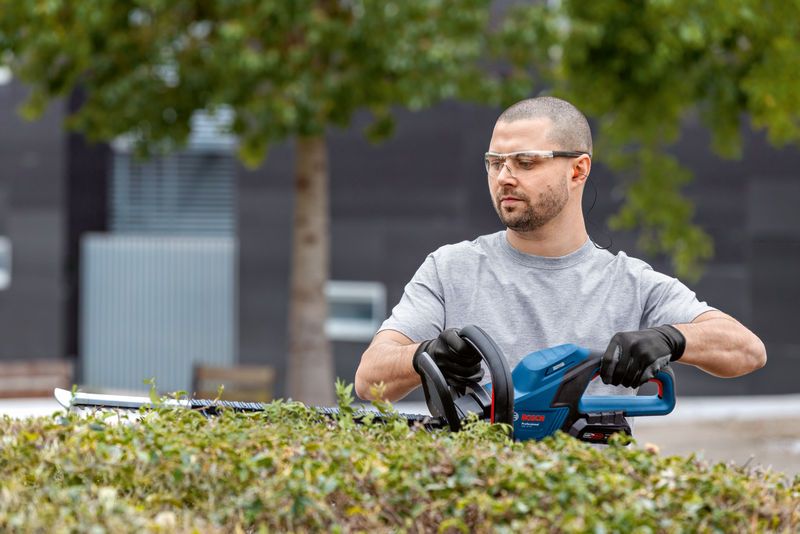 A person wearing safety equipment trims bushes using a cordless hedge trimmer.