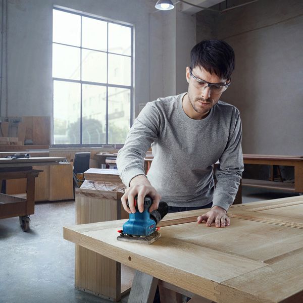 A person wearing safety equipment sands a wooden door with a handheld power sander.