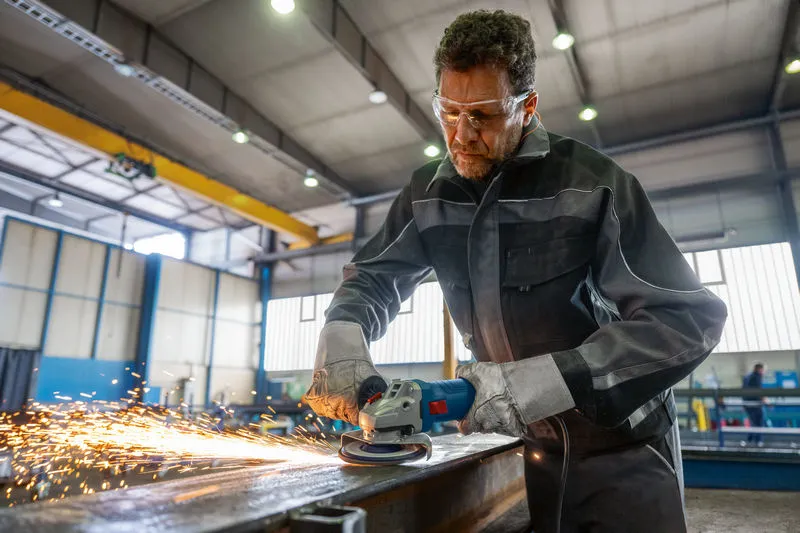 A person wearing safety equipment grinds metal, producing sparks in an industrial workshop.