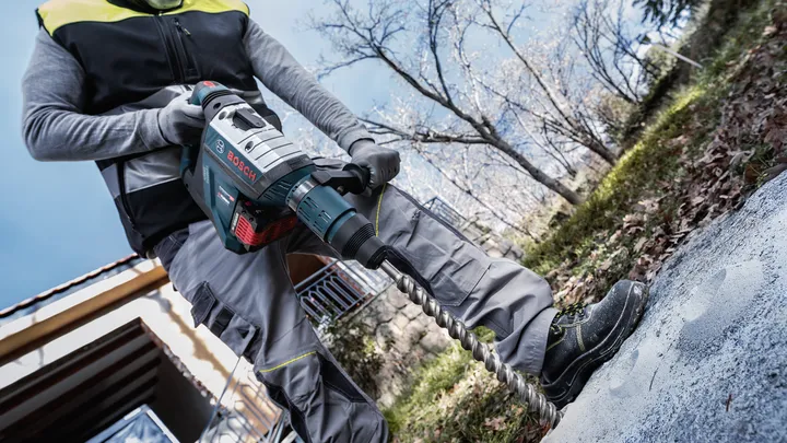 A person wearing safety equipment drills into concrete outdoors.