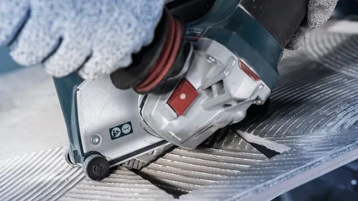 Person wearing safety equipment operates a power tool to cut a grooved metal surface.