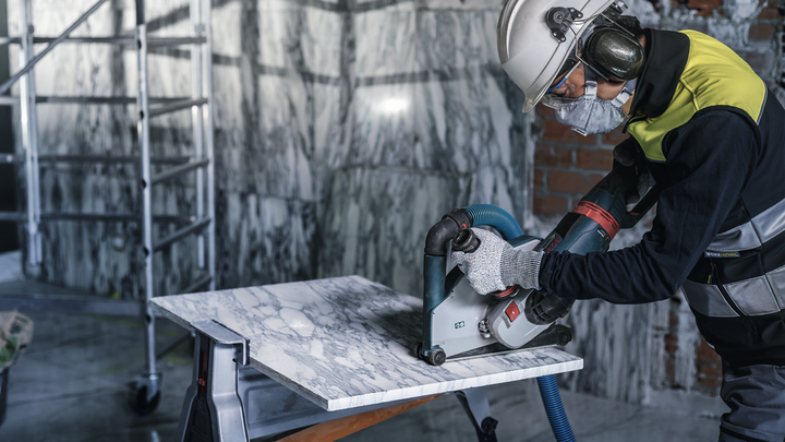 A worker wearing safety equipment cuts a marble slab with a power saw.