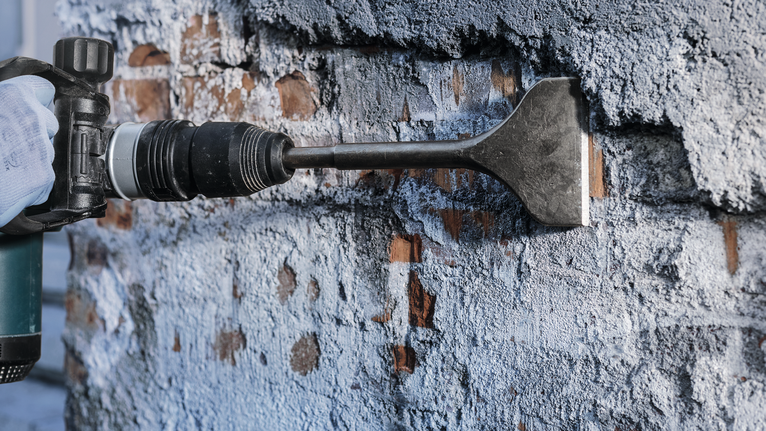 Person wearing safety equipment uses a chisel attachment to remove plaster from a wall.