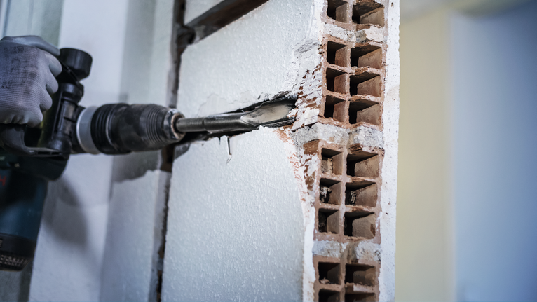 A person wearing safety equipment uses a rotary hammer to chip away a brick wall.