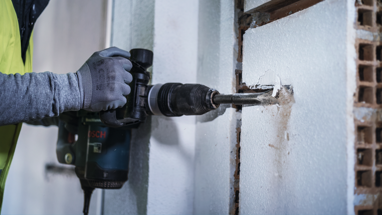 A person wearing safety equipment uses a power drill to bore a hole in a wall.