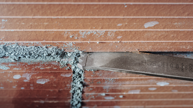 Chisel removing mortar between brown tile joints.