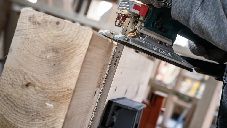 Person wearing safety equipment cuts a thick wooden plank with a jigsaw.