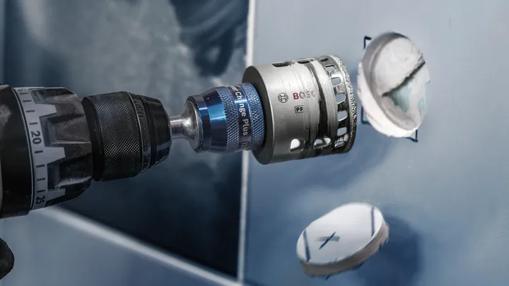 A hole saw attached to a drill creates a circular opening in blue tile.