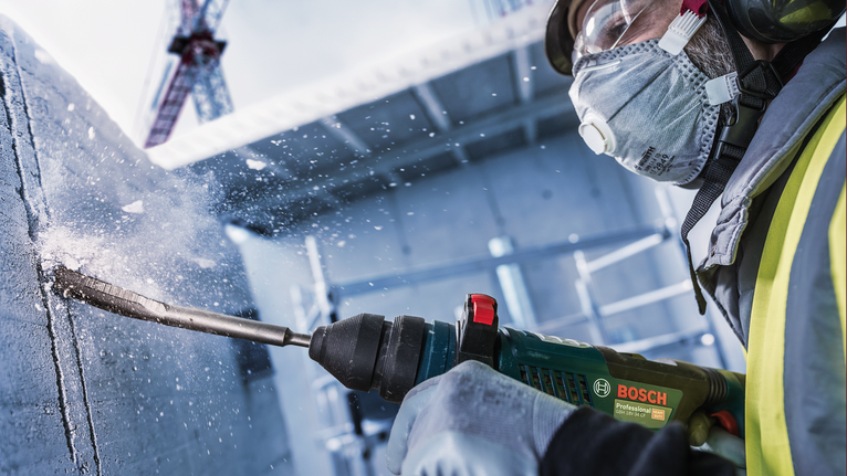 A person wearing safety equipment drills into concrete, creating flying debris.