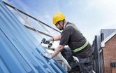 A person wearing safety equipment drives a screw into a metal roof with a cordless drill.