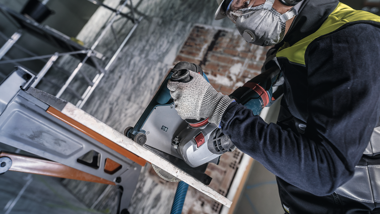 Person wearing safety equipment drills into a concrete wall with a power tool.