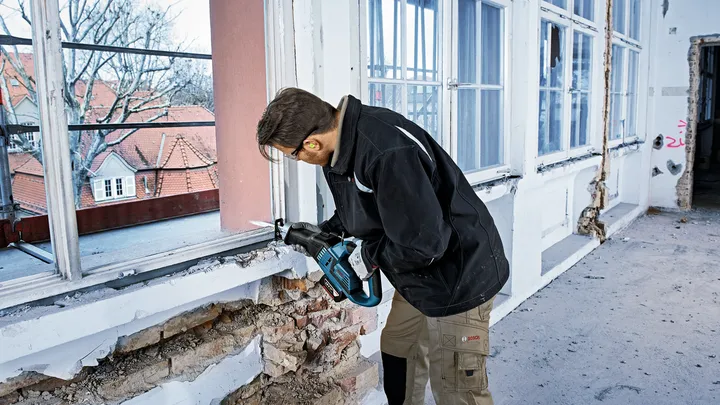 Person wearing safety equipment uses a power tool to cut into a damaged window frame.