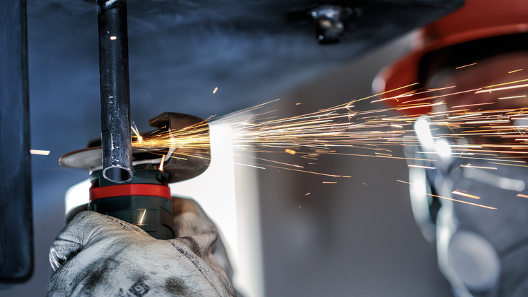 Person wearing safety equipment grinds metal pipe as sparks fly.