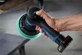 A person holds a cordless polisher with a green foam pad near a workbench.
