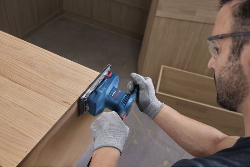 A person wearing safety equipment sands a wooden surface with an orbital sander.