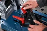 A person adjusts a metal cut-off saw angle, shavings cover the blue base.