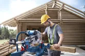 A person wearing safety equipment cuts wood with a mitre saw at a log house construction site.