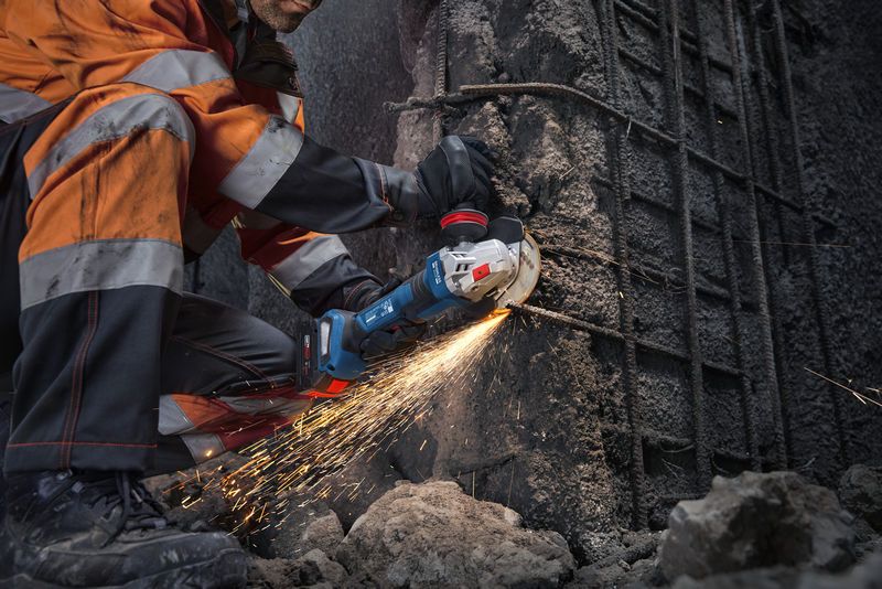 A person wearing safety equipment cuts concrete with an angle grinder, producing sparks.
