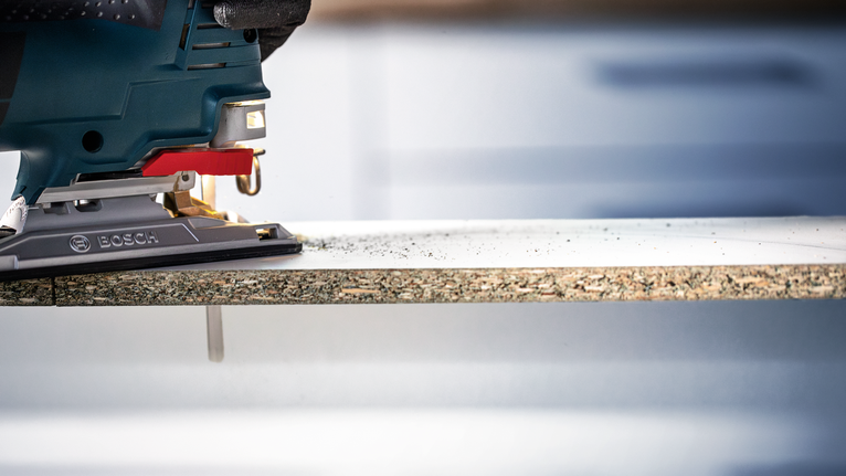Person wearing safety equipment uses a jigsaw to cut a laminated board.